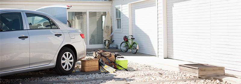 A car parked in front of a house with its trunk open. Piled behind the car are some items indicating the owner is going on a trip to the lake: a pair of flippers, a picnic basket, two fishing poles, and a cooler.