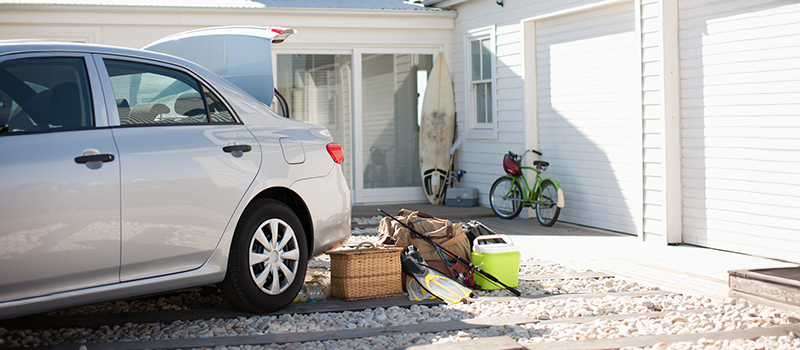 A car parked in front of a house with its trunk open. Piled behind the car are some items indicating the owner is going on a trip to the lake: a pair of flippers, a picnic basket, two fishing poles, and a cooler.